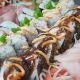 Elegant presentation of fresh sushi rolls featuring creative garnishing with spicy mayo and vibrant tobiko. The diagonal composition leads the eye through various textures and preparations, from maki to fresh sashimi.