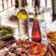 A casual yet sophisticated spread featuring a fresh-baked pizza with premium toppings, paired with Ménage à Trois white wine. The warm lighting and rustic table setting create an inviting Mediterranean atmosphere.